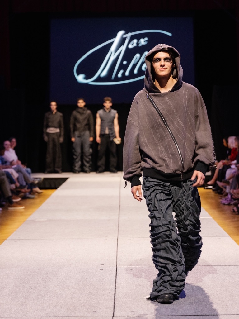 Runway photograph of Below the Surface look 1: an asymmetric taupe hooded top with densely gathered charcoal trousers, front walk