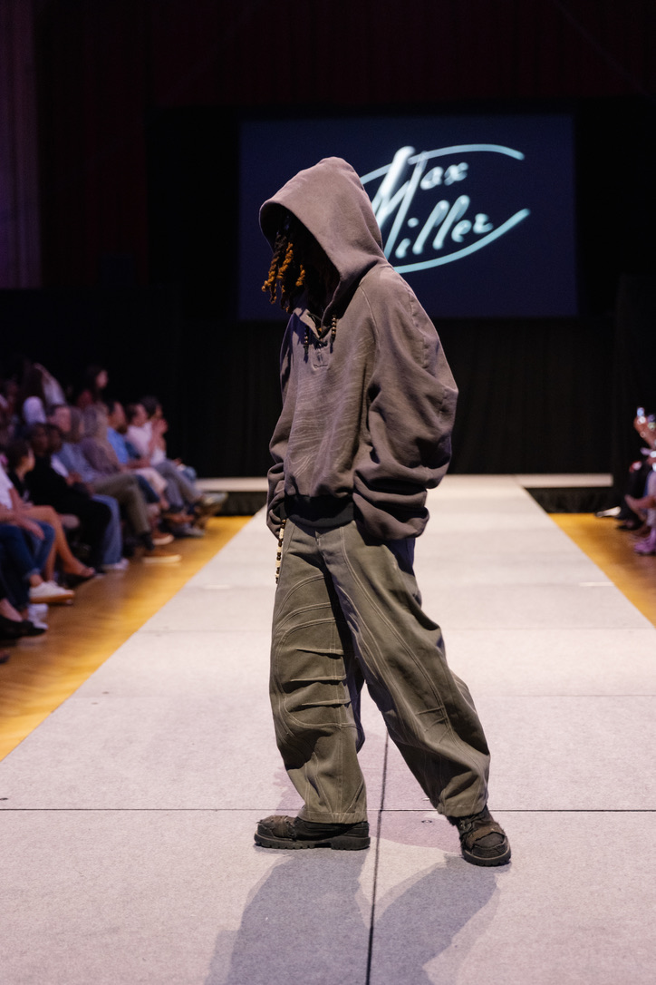 Runway photograph of Below the Surface look 2: a soft taupe hooded top with olive engineered trousers, side profile