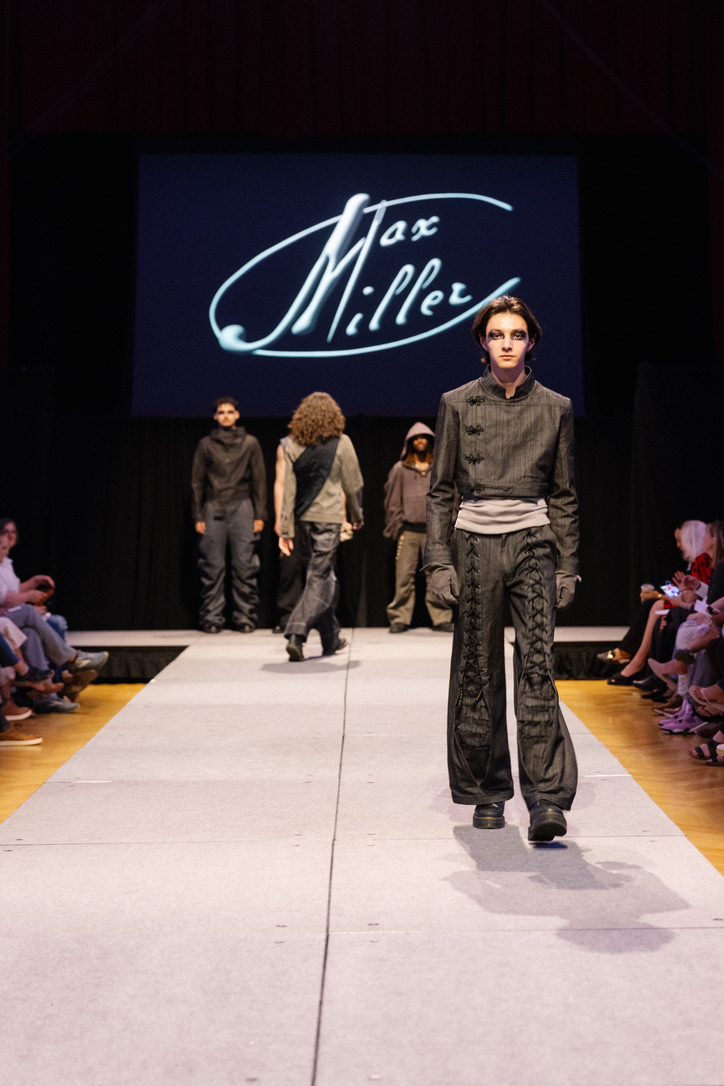 Runway photograph of Below the Surface look 3: a cropped charcoal jacket with frog closures and laced trousers, front walk