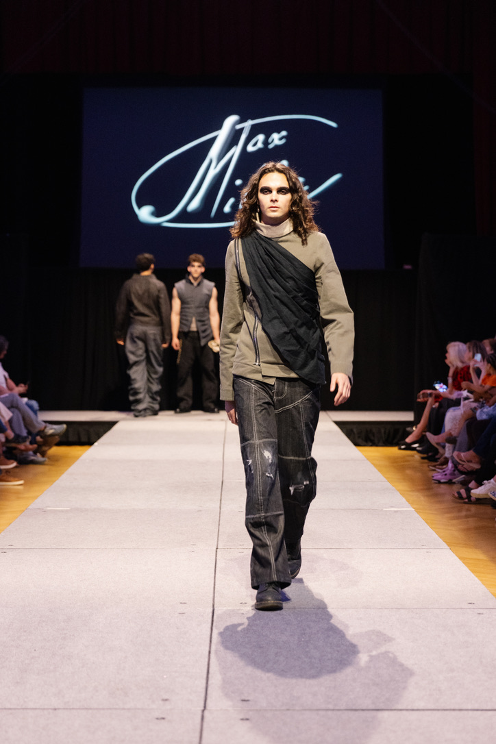Runway photograph of Below the Surface look 4: an olive jacket crossed by a draped black panel with dark paneled trousers, front walk