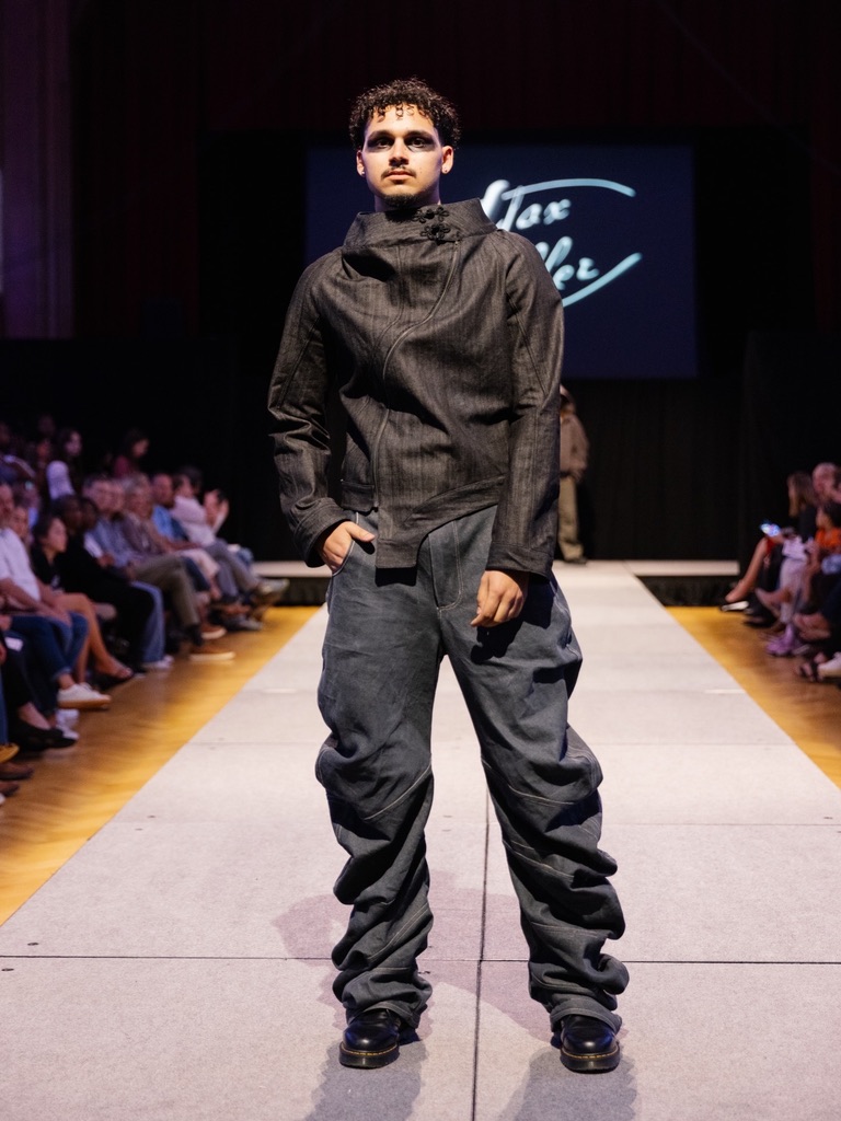 Runway photograph of Below the Surface look 5: a dark asymmetric raised-collar jacket with gray gathered trousers, front stance