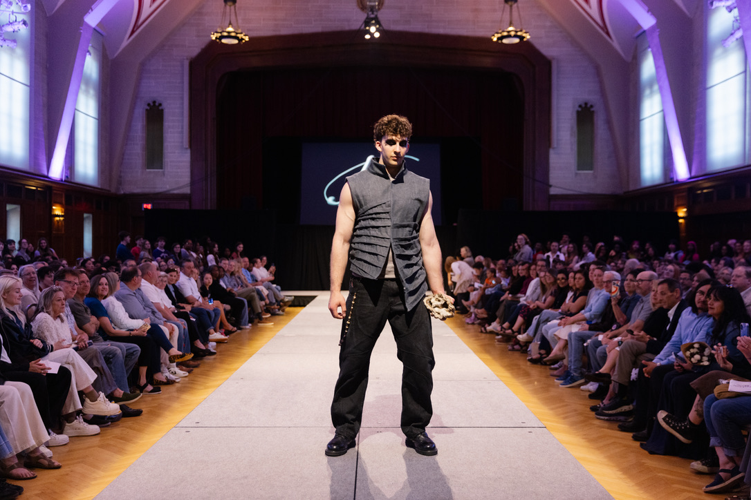 Runway photograph of Below the Surface look 6: a sculptural sleeveless charcoal vest with dark engineered trousers, front stance, landscape